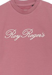 T-shirt in cotone rosa chiaro con testo ricamato bianco "Roy Roger's." Collo rotondo con bottoni su una spalla per una facile vestibilità.
