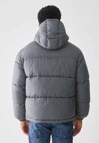 Chaqueta acolchada gris con capucha que presenta secciones acolchadas, puños elásticos y un acabado suave. Combinada con jeans de mezclilla azules.