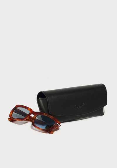 Óculos de sol de tartaruga com lentes escuras ao lado de uma case de couro preto com aba arredondada e logótipo em relevo.