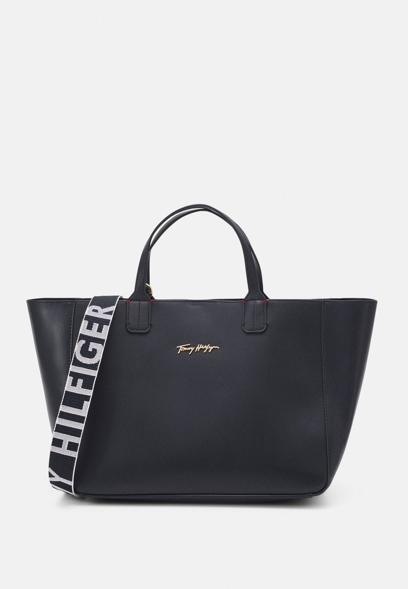 Sac tote en cuir noir avec double poignées, bandoulière en tissu de marque amovible et logo Tommy Hilfiger en métal doré à l'avant.