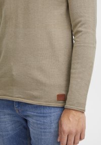 Haut à manches longues en tricot marron clair avec un motif texturé et une étiquette de marque en cuir marron sur l'ourlet inférieur. Porté avec un jean en denim bleu.