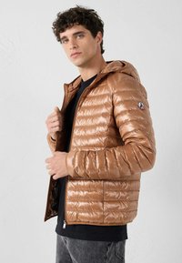 Giacca imbottita marrone con cappuccio, finitura lucida, polsini a coste e chiusura frontale con zip. Patch logo sul braccio sinistro.