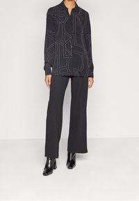 Camicia nera con bottoni e cuciture bianche geometrica, abbinata a pantaloni neri a gamba larga e stivali neri con tacco a punta.