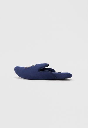 Chausson bleu marine à enfiler avec broderie florale sur le dessus, tissu doux et design ouvert à l'arrière sur fond gris clair.