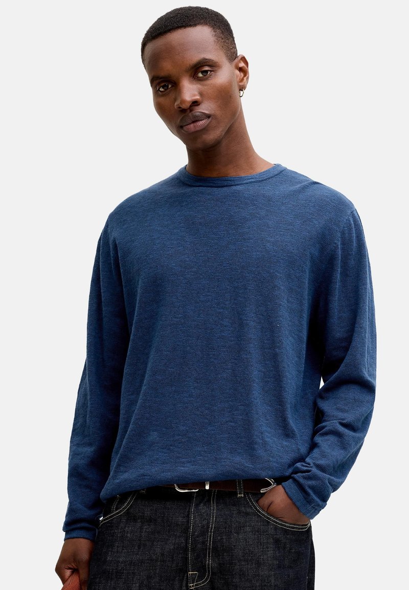 Homme portant un pull à manches longues bleu et un jean sombre, debout avec une main dans la poche sur un fond clair uni.