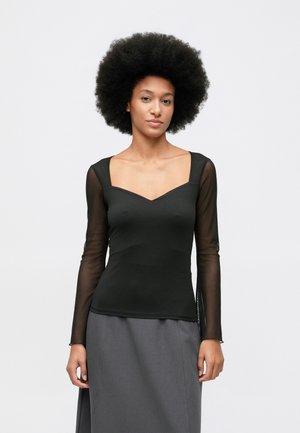 Femeie cu coafură afro naturală, purtând un top negru ajustat cu mâneci lungi transparente și o fustă gri, stând în fața unui fundal alb.