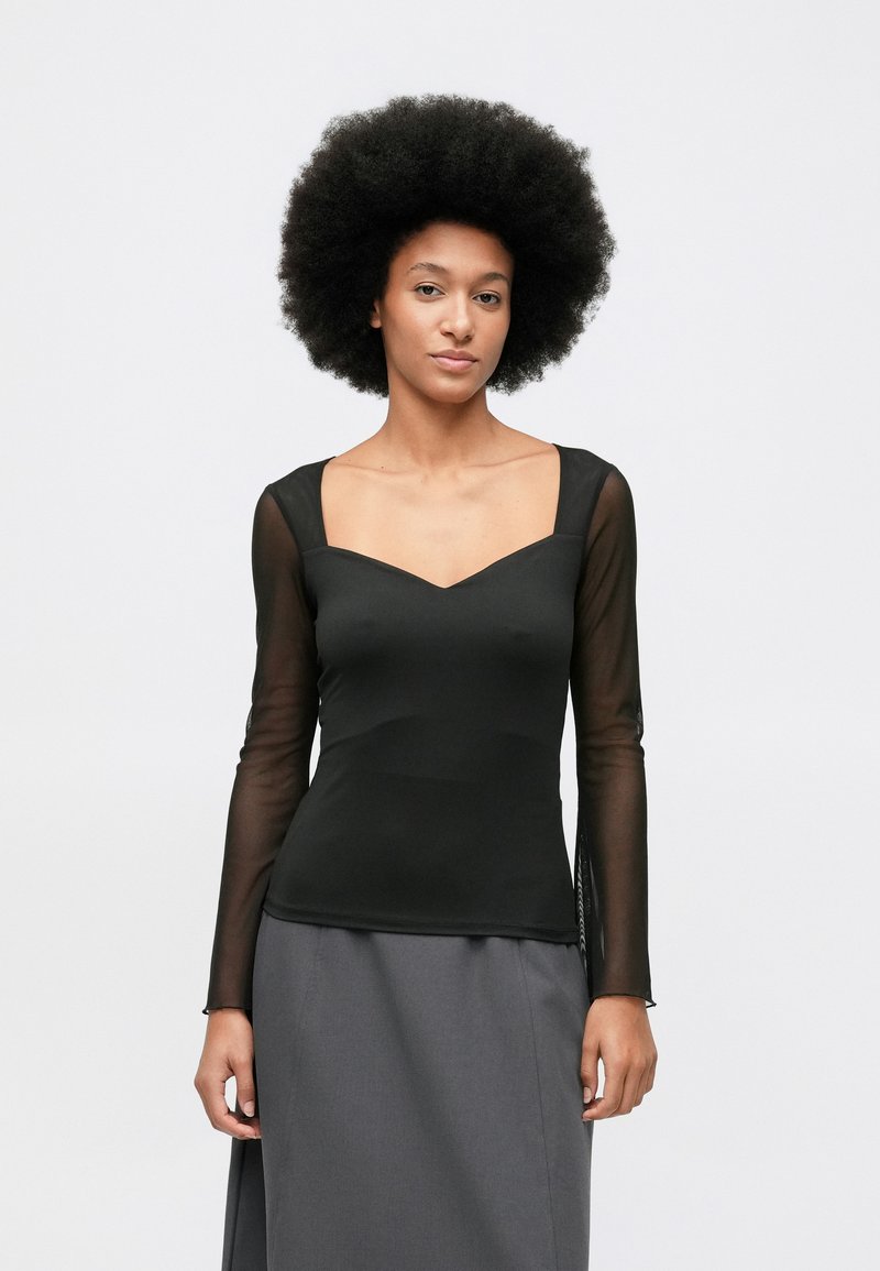 Moteris su natūraliu afro šukuosena, vilkinti juodą prigludusį marškinį su permatomomis ilgomis rankovėmis ir pilku sijonu, stovi priešais baltą foną.