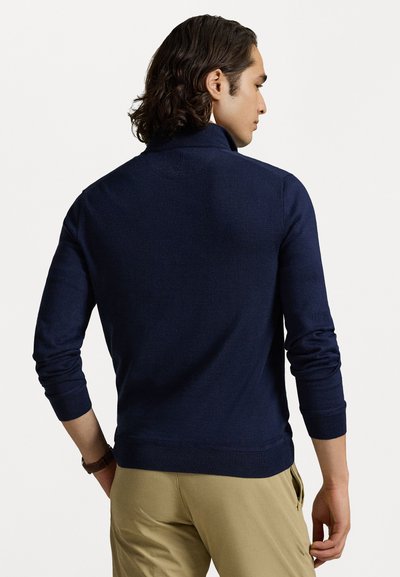 Pulover bleumarin cu guler înalt, dintr-un material tricotat texturat și cu manșete ribbed; purtat cu pantaloni khaki.