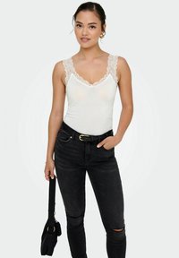 Hvid tanktop med blonder langs halsudskæringen og stropperne, parret med højtaljede mørke jeans. Sort håndtaske holdt i den ene hånd.