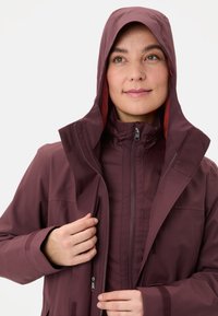 Giacca impermeabile bordeaux con cappuccio, caratterizzata da un interno rosso a contrasto, chiusura con zip e dettagli in velcro su colletto e polsini.