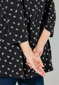 Zwarte shirt met wit bloemenpatroon, met 3/4 mouwen en een losse pasvorm. Soepele textuur, gedragen over blauwe jeans, handen achter de rug gevouwen.