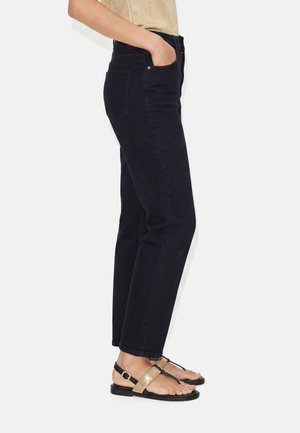 Person trägt schwarze Slim-Fit-Jeans und goldene Sandalen mit Riemchen, steht mit einer Hand in der Tasche vor neutralem Hintergrund.