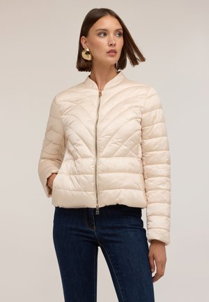 Donna che indossa una giacca trapuntata crema con cerniera e colletto rialzato, jeans blu scuro e grandi orecchini dorati, guarda di lato con una mano in tasca.
