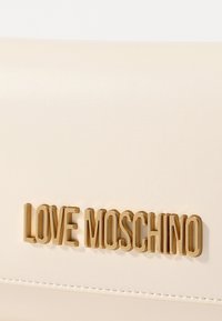 Cream-colored clutch bag made of textured material, featuring a gold "LOVE MOSCHINO" logo and visible stitching along the edges.