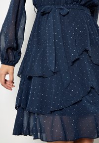 Granatowa warstwowa sukienka z lekkiego materiału, z paskiem do wiązania, długimi rękawami oraz drobnymi srebrnymi kropkami w całym fasonie.