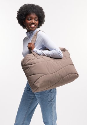 Femme souriante portant un pull-over bleu clair et un jean, tenant un grand sac fourre-tout en cuir beige sur l'épaule, devant un fond blanc.