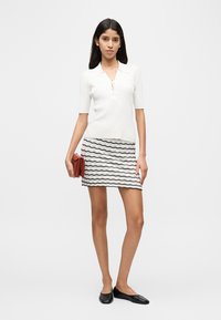 Άσπρο ριγωτό τοπ με κοντά μανίκια και φερμουάρ στον γιακά, σε συνδυασμό με μίνι φούστα με μοτίβο κυματιστών μαύρων γραμμών και κόκκινο πορτοφόλι. Μαύρες μπαλαρίνες.