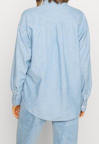 Jasnoniebieska koszula z denimu z kołnierzykiem zapinanym na guziki, długimi rękawami i luźnym krojem. Tył koszuli posiada detal w postaci yoku i plisę.