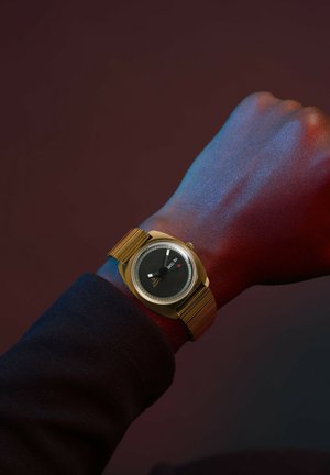 Zlatý náramkový hodin s kulatým černým ciferníkem, bílými hodinovými značkami a kovovým páskem na ruce na tmavém pozadí.