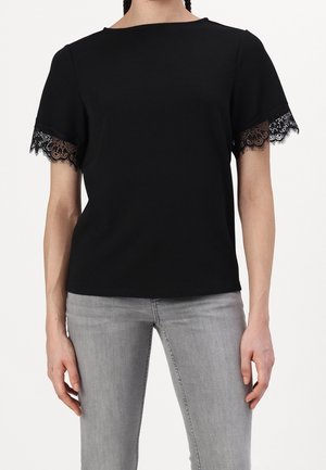 Kobieta ubrana w czarny krótki top z koronkową lamówką na rękawach oraz jasnoszare obcisłe dżinsy, stojąca na jednolitym tle.