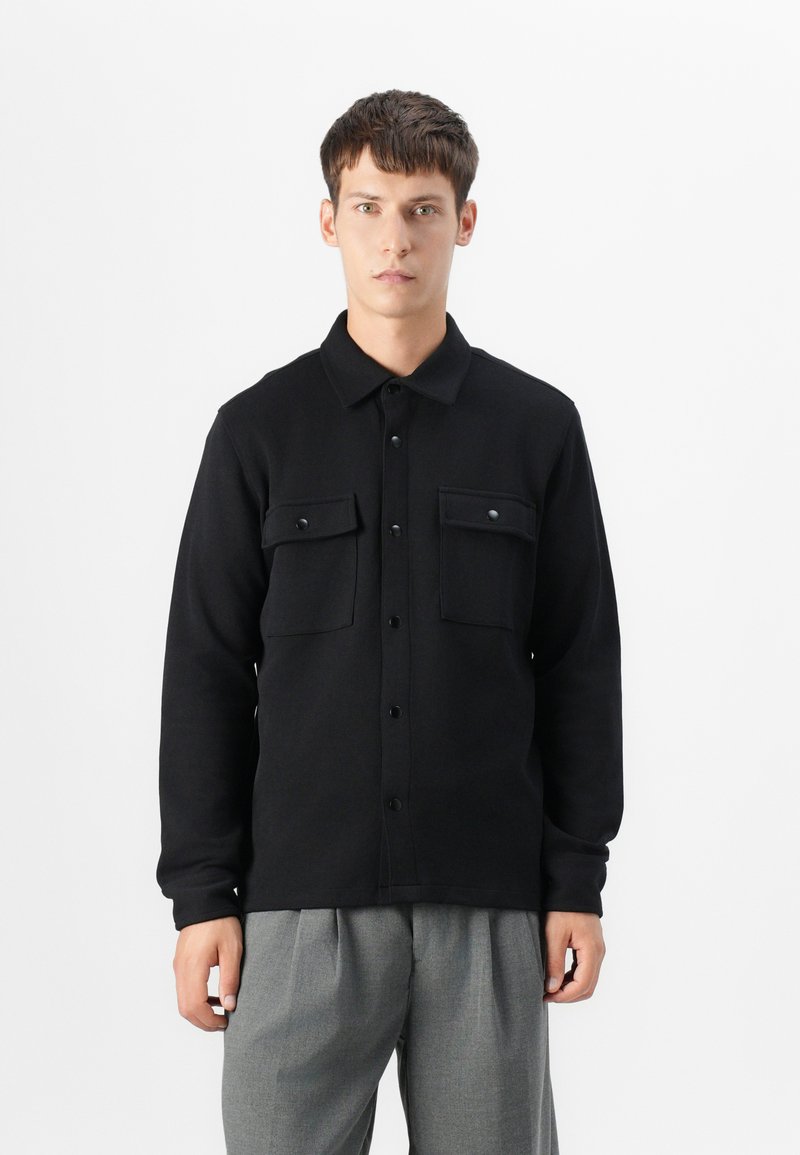 Black long-sleeve button-up shirt with two chest pockets, crafted from textured fabric, featuring a collared neck and front button closure.