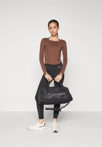 Rjav dolg rokav s črnimi šivi, črne leggins hlače in črna torba za šport. Model nosi bele superge s rjavimi dodatki.