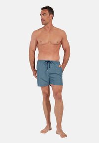 Homme musclé debout pieds nus, portant un short de bain bleu à motifs avec un cordon noir, regardant vers sa gauche.