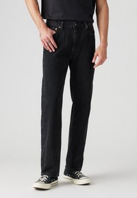 Homme portant un jean noir droit, un t-shirt noir et des baskets noires et blanches, debout avec les mains détendues le long du corps.