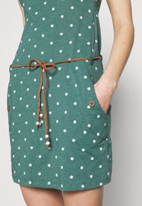 Robe turquoise à pois blancs, dotée de deux poches latérales, d'une ceinture tressée marron avec des perles et d'une coupe décontractée.