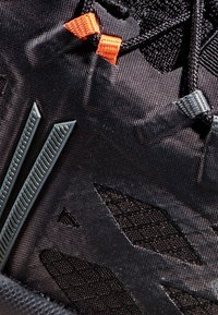 Schwarzer Sportschuhbereich mit strukturiertem Mesh, Gummielementen und kontrastierenden orangefarbenen und grauen Schnürsenkeln. Enthält anguläre Designelemente.