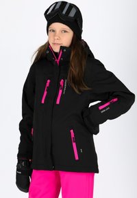 Giacca invernale nera con cerniere rosa, numerose tasche e polsini regolabili. Indossata con pantaloni rosa e un berretto nero con occhiali da sci.