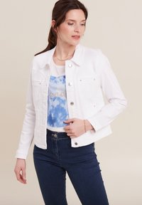 Witte denimjacket met knoopsluiting, twee voorzakken en zilverkleurige hardware. Gekozen boven een blauwe grafische top en donkere jeans.
