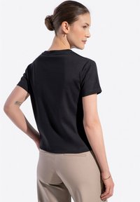 T-shirt nera in cotone con maniche corte, scollatura rotonda, mostrata dal retro, abbinata a pantaloni beige. Tessuto liscio, design minimal.