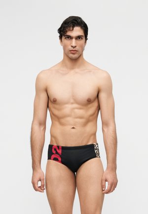 Mladý muž s tmavými vlasmi má na sebe čierne plavecké slipy s červeným nápisom "D2" a leopardím vzorom na paneli, stojaci pred jednofarebným svetlým pozadím.