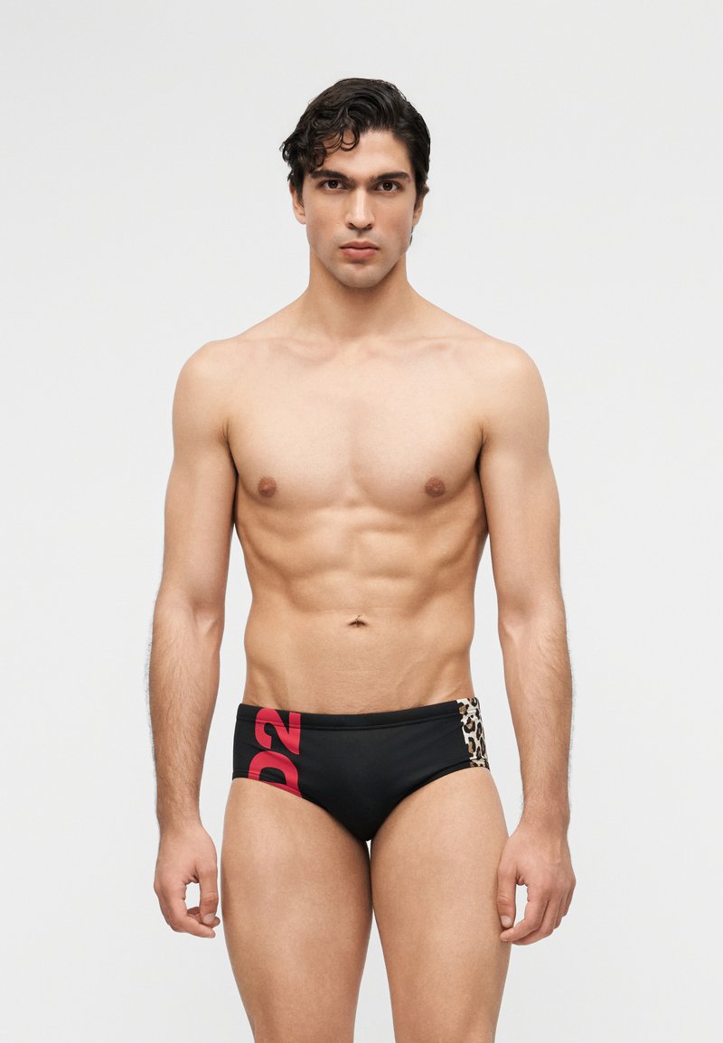 Νεαρός άνδρας με σκούρα μαλλιά φοράει μαύρα μαγιό με κόκκινο κείμενο "D2" και πάνελ λεοπάρ, στέκεται μπροστά σε απλό ανοιχτό φόντο.