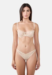 Soutien-gorge en dentelle avec armatures et string assorti en beige avec des motifs floraux, des bretelles fines et des bords festonnés, comportant un détail en trou de serrure au centre.