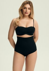 Blondynka w czarnych majtkach z wysokim stanem i strapless biustonoszu stoi na tle jasnym, zwrócona przodem z rękami za plecami.