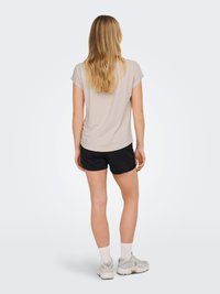 Femme aux longs cheveux blonds, vue de dos, portant un haut beige à manches courtes, un short noir, des chaussettes blanches et des baskets de couleur claire sur fond blanc.