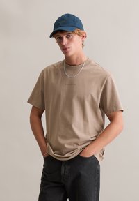 Beżowy bawełniany t-shirt z krótkimi rękawami, okrągłym dekoltem i małym czarnym logo, zestawiony z ciemnymi dżinsami i niebieską czapką z daszkiem.