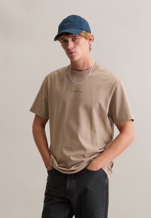 Beżowy bawełniany t-shirt z krótkimi rękawami, okrągłym dekoltem i małym czarnym logo, zestawiony z ciemnymi dżinsami i niebieską czapką z daszkiem.