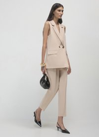 Gilet beige doppiopetto con due tasche frontali, abbinato a pantaloni affusolati coordinati. Completano il look una borsa nera lucida e décolleté con punta.
