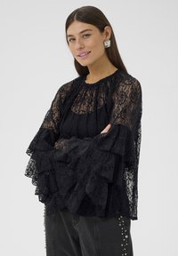 Melna mežģīņu blūze ar volānu piedurknēm, augstu apkakli, sarežģītu ziedu rakstu un daudzslāņu tekstūrām. Kombinēta ar tumšām džinsa biksēm.