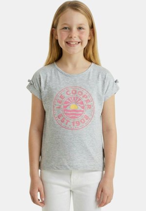 Fille souriante aux longs cheveux blonds portant un T-shirt gris avec un logo circulaire rose Lee Cooper et un pantalon blanc, debout devant un fond blanc.