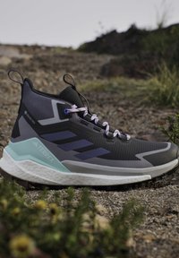 Chaussure de trail running extérieure noire et grise avec des touches bleu clair, étiquette GORE-TEX et semelle texturée sur fond de terrain rocheux.