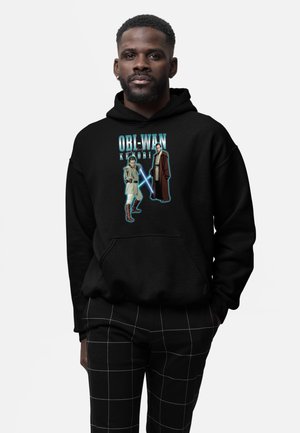 Mand iført sort hættetrøje med Obi-Wan Kenobi-grafik, der viser to Jedi med lyssværd, og sorte bukser med hvidt gittermønster.