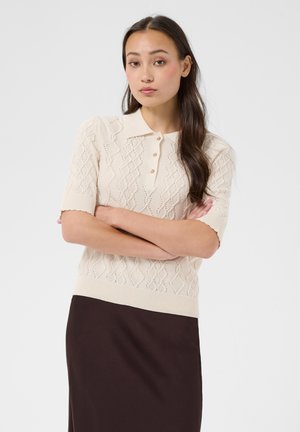 Jeune femme aux longs cheveux foncés portant un haut court en maille crème avec des boutons et une jupe marron foncé, debout les bras croisés.
