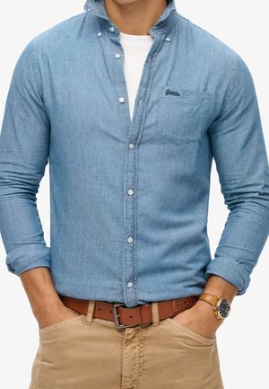 Mann trägt hellblaues Hemd über weißem T-Shirt, beige Hose mit braunem Gürtel, Hände in den Taschen und goldene Armbanduhr am linken Handgelenk.