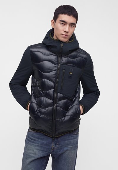Jeune homme portant une veste matelassée noire avec capuche et poche poitrine zippée, mains dans les poches latérales, debout devant un fond uni.