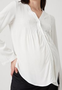 Vit gravidblus med långa ärmar, plisserad frontdetalj, v-ringning och knappstängning, tillverkad av lättviktig tyg.