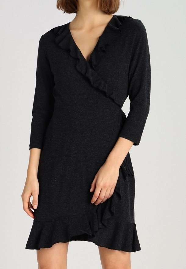 Femme portant une robe portefeuille gris foncé avec bordure à volants et manches trois-quarts sur un fond neutre.